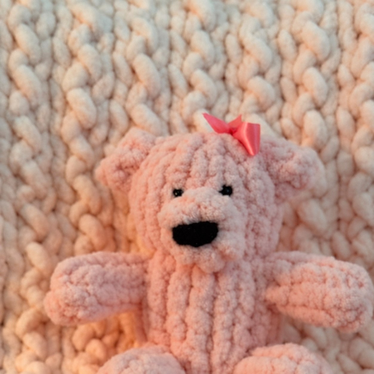 Pink handmade teddy bear resting on a chunky knit blanket in cream, pale blue, and pink